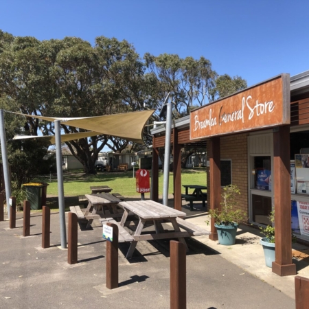 the outside of the Breamlea General store, including tables, wooden bollards, trees and grass and a blue sky